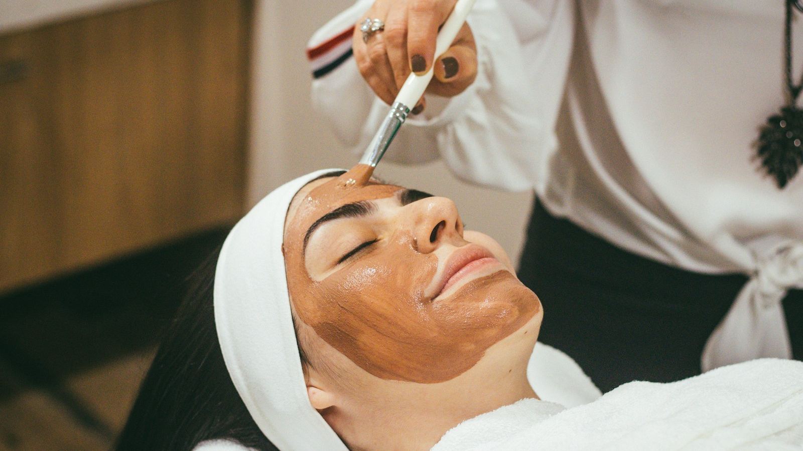 A woman lying back with her eyes closed while a therapist brushes a facial mask onto her skin.