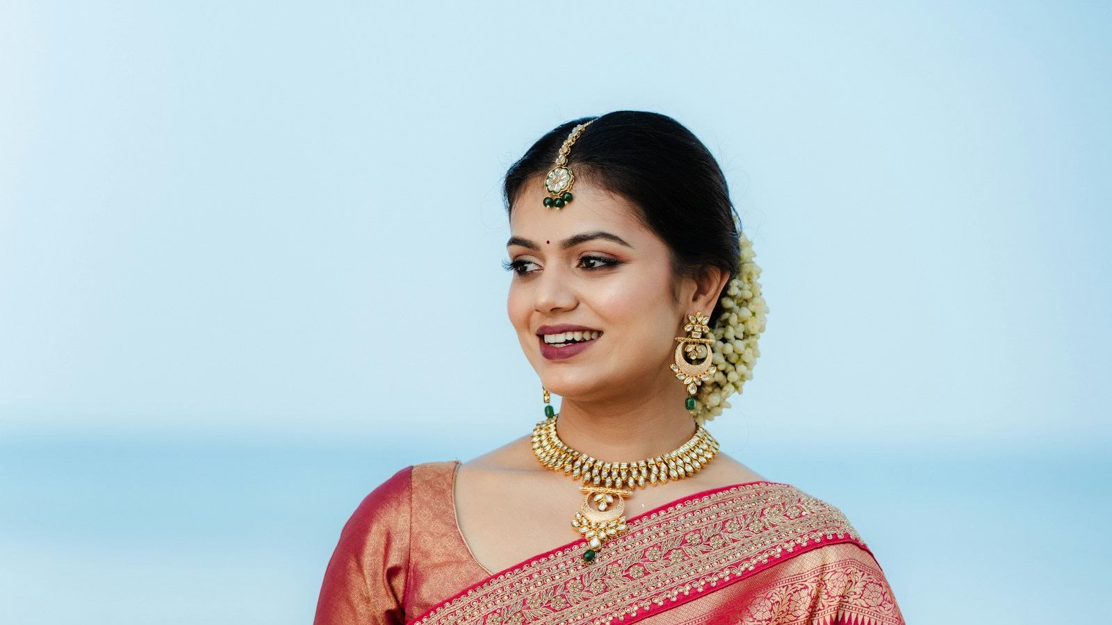 A smiling woman in a red and gold silk saree, with soft, natural-looking makeup and jasmine in her hair.