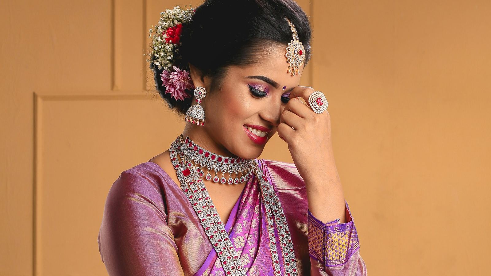 A woman in a purple and gold silk saree with flowers in her hair, smiling and looking down.