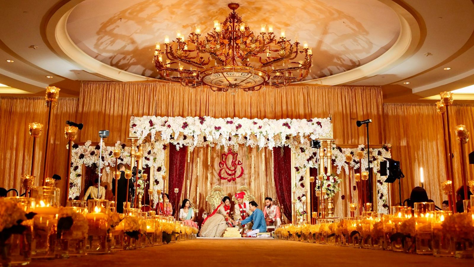A candle-lit wedding hall with a flower-covered mandap and the couple seated at its centre.